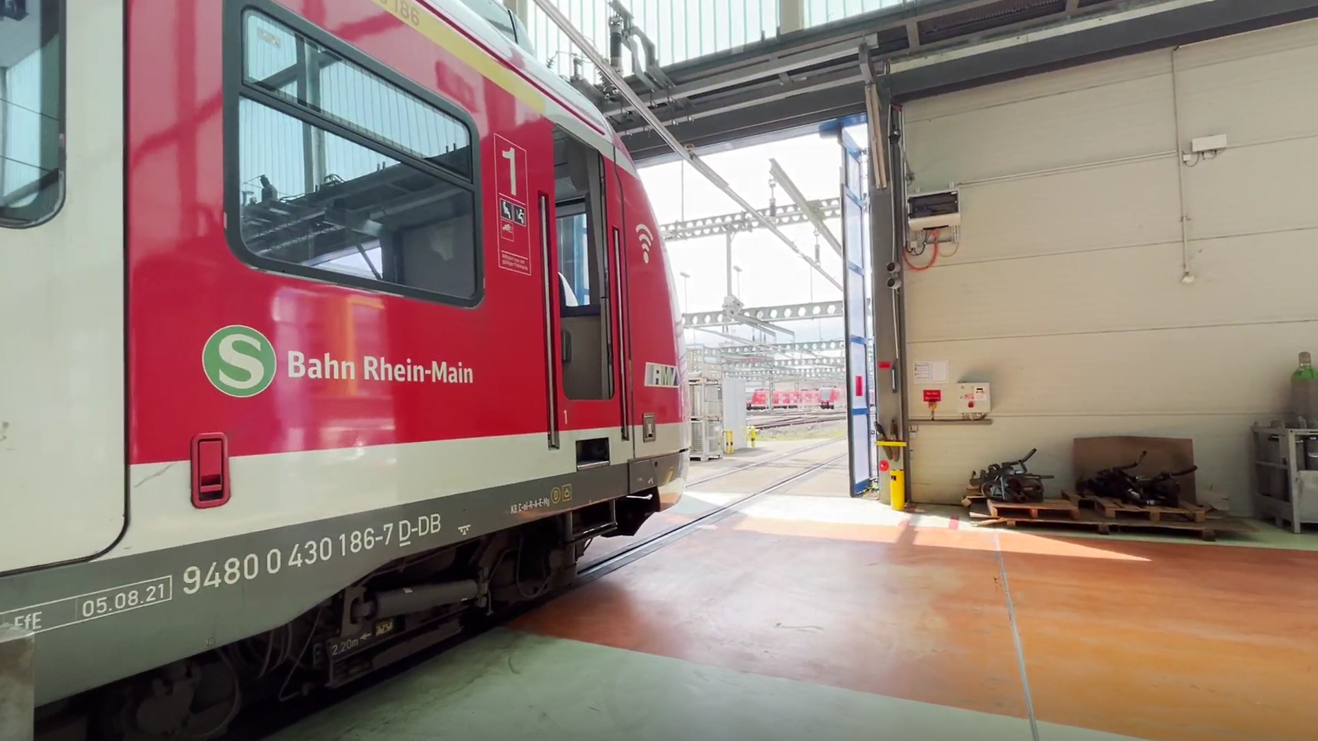 Ein roter Zug steht auf Gleisen in in der Halle eines Instandhaltungswerks, die Hallentore schließen sich hinter ihm. 