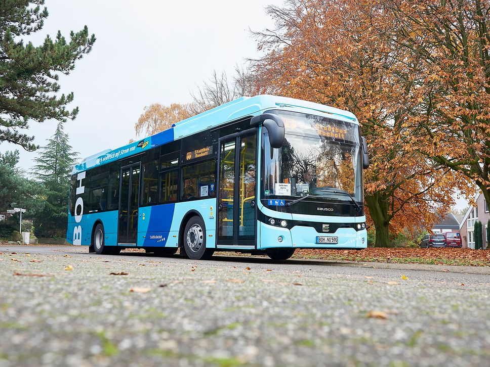 A light blue bus stands on a road with autumnal trees in the background