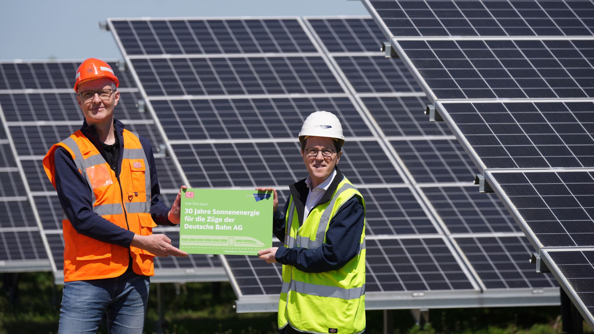 Two men in construction helmets and high-visibility vests hold an advertising sign and stand in front of a PV system.